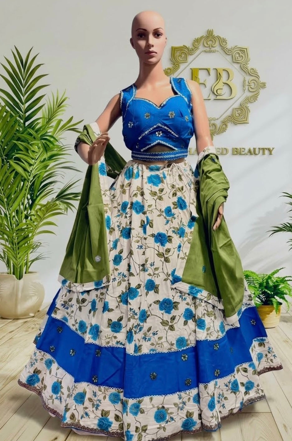 A womannequin displays a traditional Indian ethnic outfit consisting of a blue embroidered sleeveless blouse and a tiered cream skirt with blue floral prints and green dupatta draped on both sides. The background features a light-colored wall with a gold 'EB Beauty' logo and potted green plants.