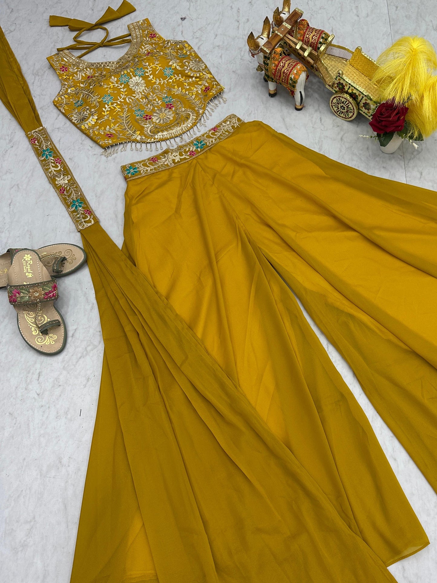 Mustard yellow traditional outfit with embroidered top, saree, and accessories on a light gray background.