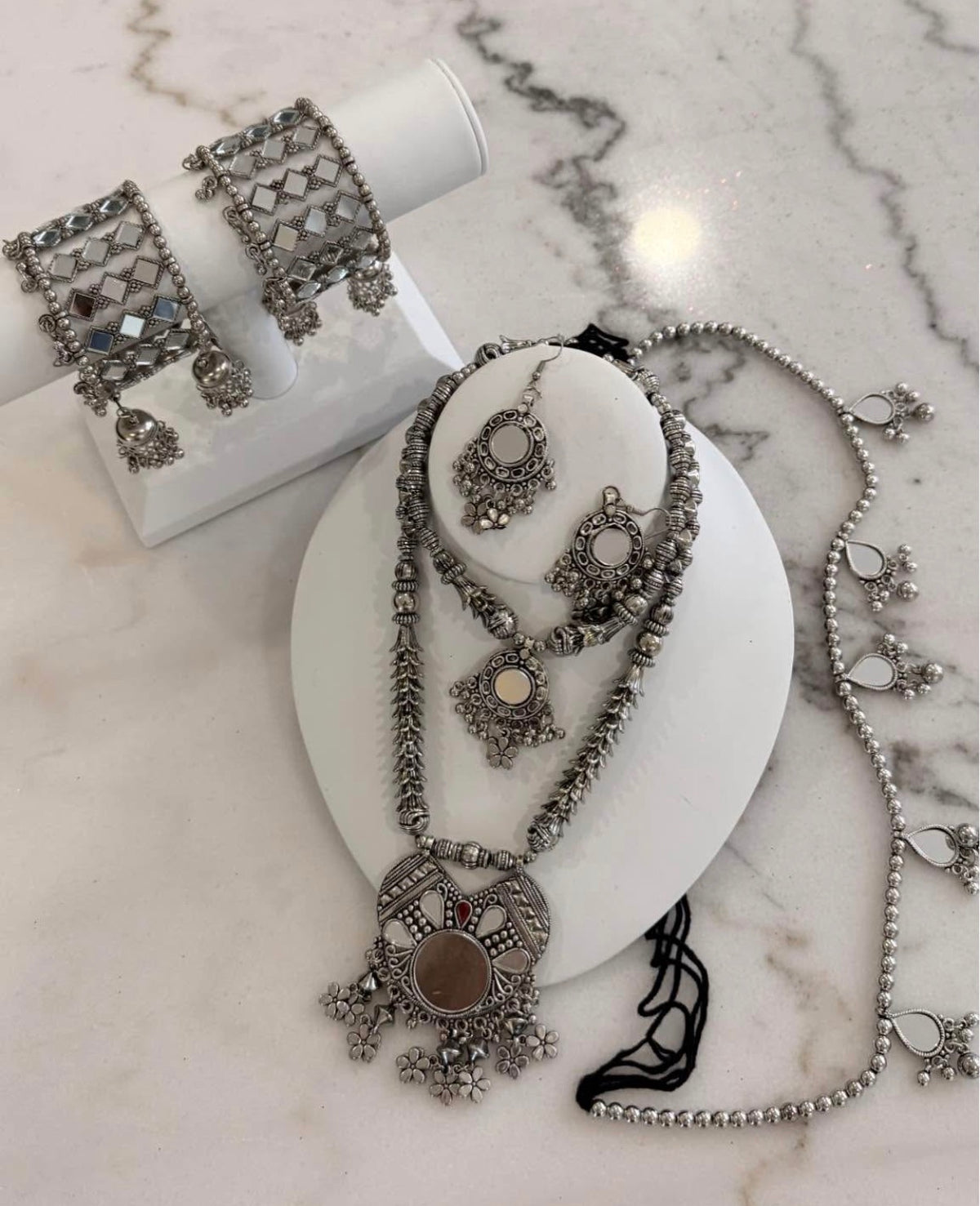 A silver-toned traditional jewelry set displayed on white mannequin stands, including a necklace with a large circular pendant, matching earrings, and a bracelet, arranged on a light marble surface.