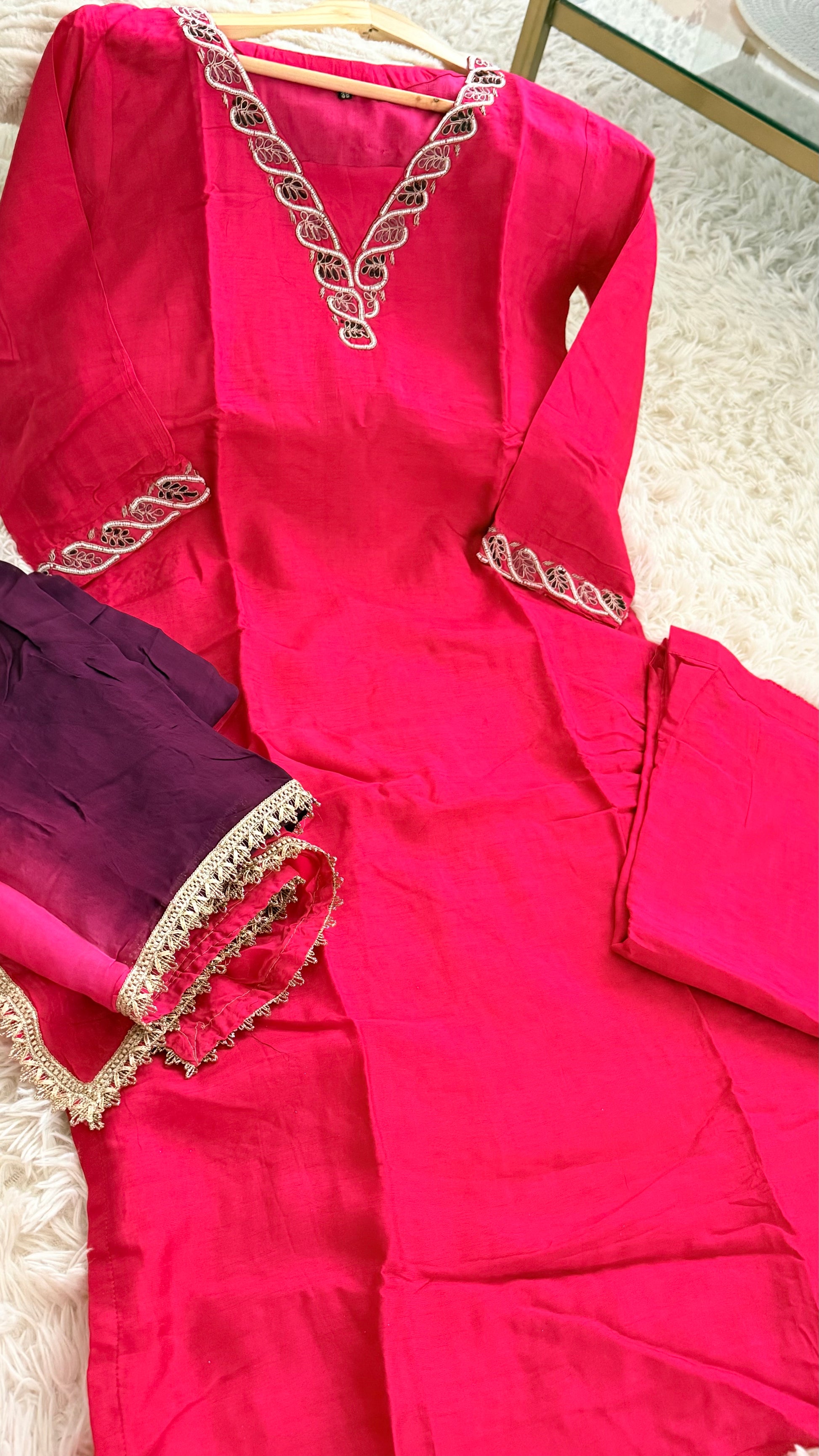 A bright pink traditional long kurta set with gold embroidered trim, laid flat on a white textured rug alongside a folded purple fabric piece with matching gold trim.