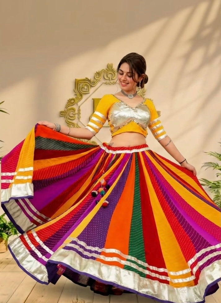 A woman modeling a vibrant, multicolored traditional Indian lehenga choli set. The skirt features bold vertical stripes of red, orange, yellow, green, purple, and white, with a matching yellow and silver choli and striped sleeves, set against a warm beige wall with decorative gold molding.