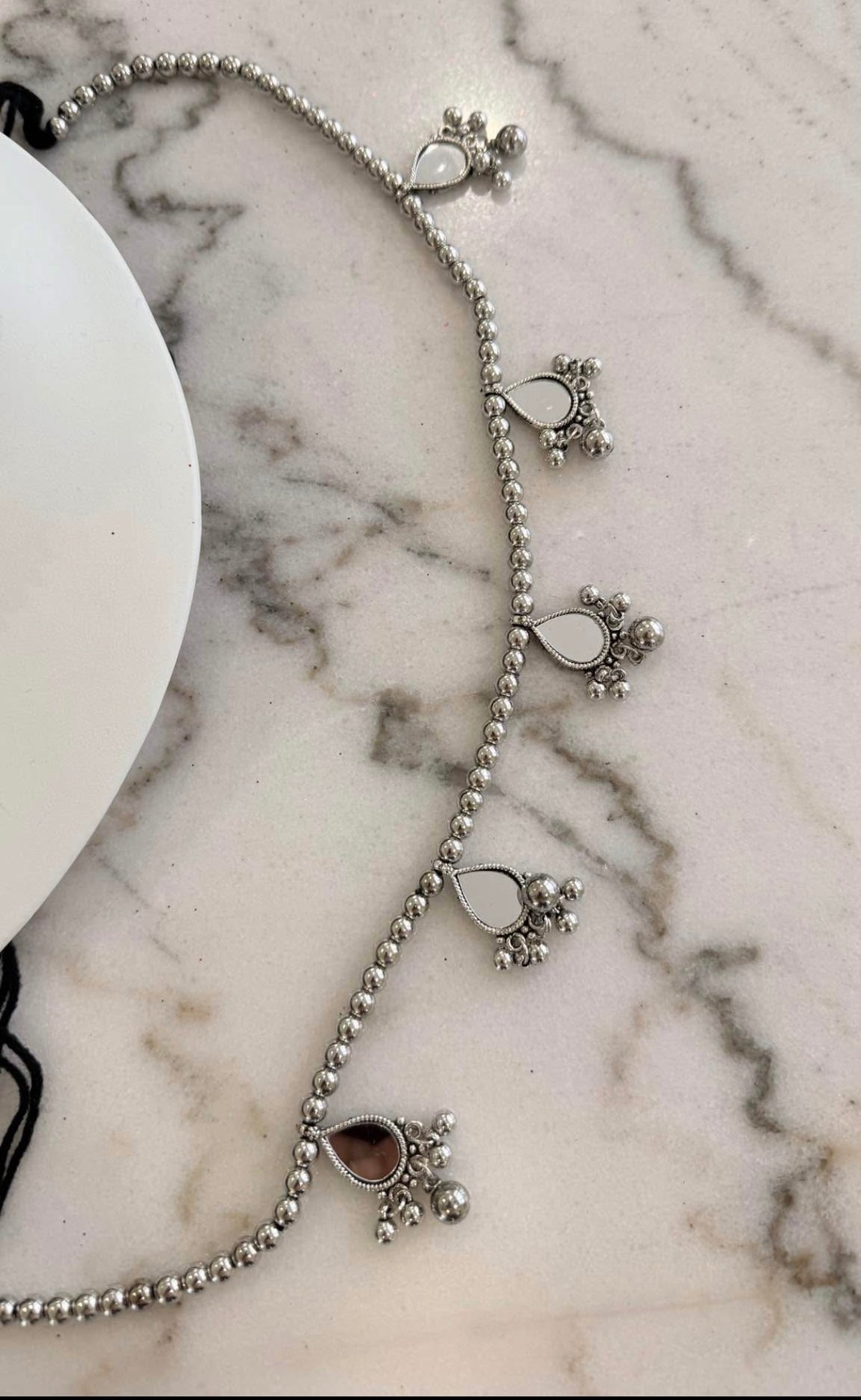 A silver beaded anklet with teardrop-shaped stone charms and small floral clusters, displayed on a white marble surface next to a white plate.