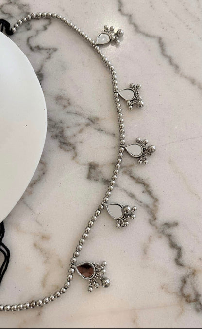 A silver beaded anklet with teardrop-shaped stone charms and small floral clusters, displayed on a white marble surface next to a white plate.