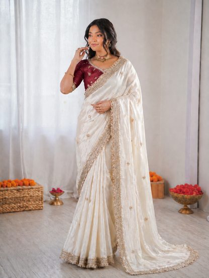 Woman wearing a white saree with gold border in a decorated room.