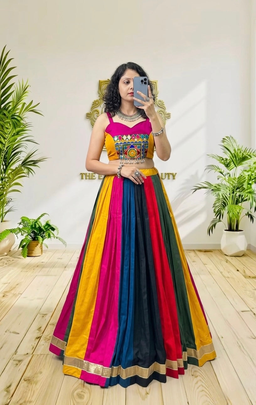 A woman takes a mirror selfie wearing a vibrant, multicolored traditional Indian lehenga choli dress. The dress features vertical panels of yellow, pink, red, and navy blue with gold borders, and a heavily embroidered yellow choli.