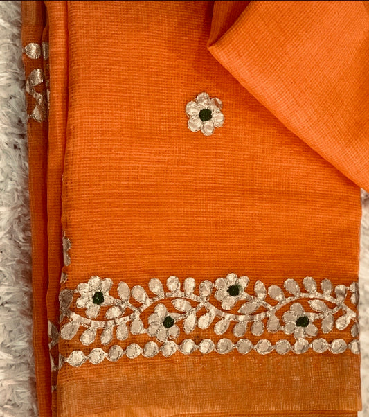 Orange fabric with a decorative border featuring silver floral embroidery and small dark green centers, shown folded on a white textured surface.