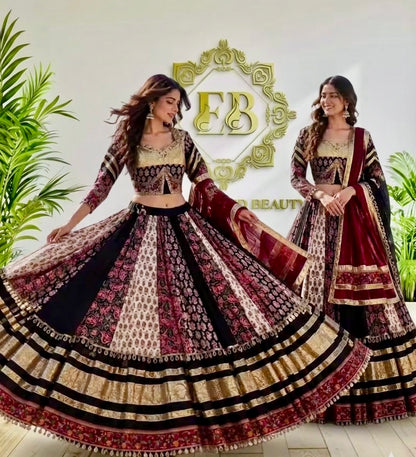 Two women modeling traditional Indian lehenga choli suits in a studio setting with decorative plants and a gold 'EB Beauty' sign on the wall. The outfits feature intricate patterns in black, beige, red, and gold.