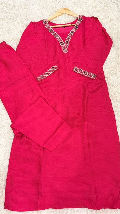 A bright pink long kurta with matching pants laid flat on a white faux fur surface. The kurta features a V-neckline and two front pockets, both detailed with metallic gold and silver chain-link embroidery.