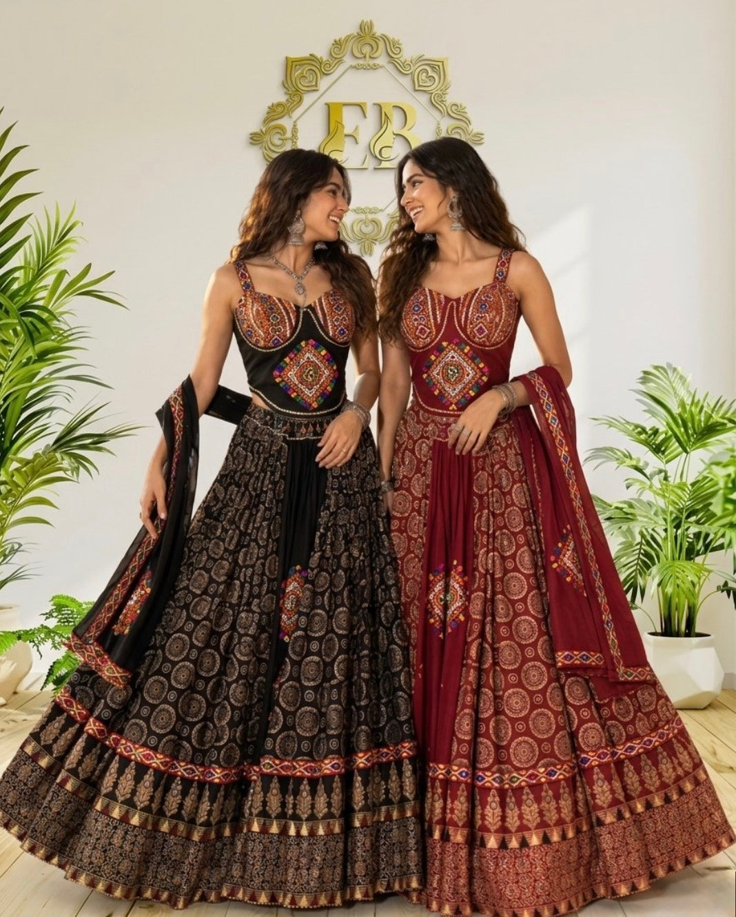 Two women modeling traditional Indian lehenga choli suits in a decorated indoor setting. One dress is black with gold and red embroidery, and the other is maroon with gold and red embroidery.