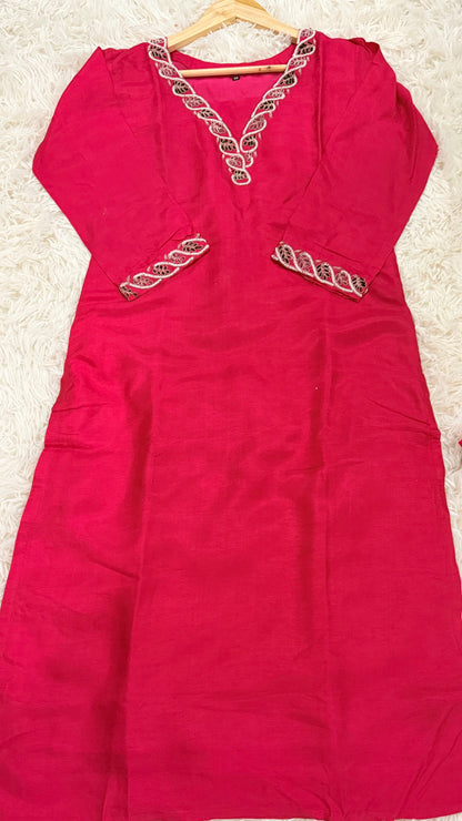 A bright pink kaftan dress with silver and gold embroidered detailing on the V-neckline and pockets, laid flat on a white shag rug.