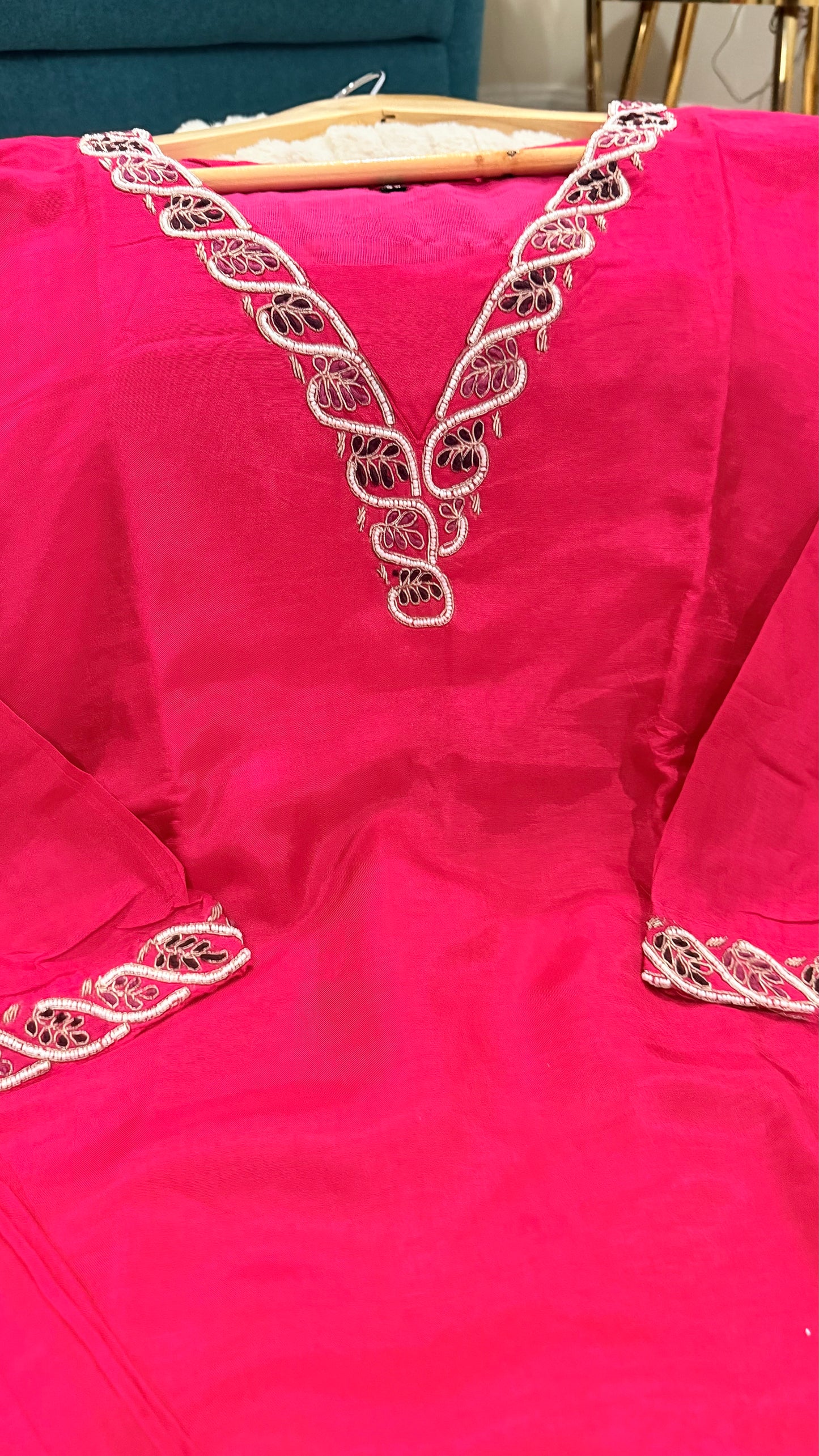 A bright pink traditional dress with intricate white and black embroidered detailing around the V-neckline and sleeve cuffs, shown hanging on a wooden hanger.