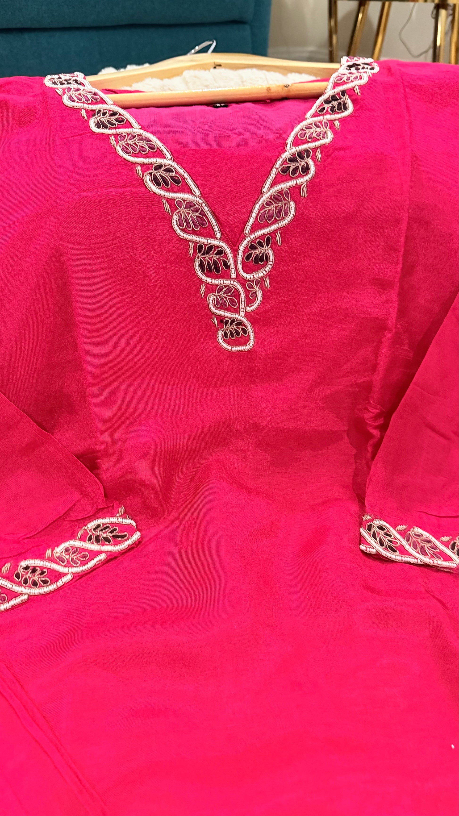 A bright pink traditional dress with intricate white and black embroidered detailing around the V-neckline and sleeve cuffs, shown hanging on a wooden hanger.