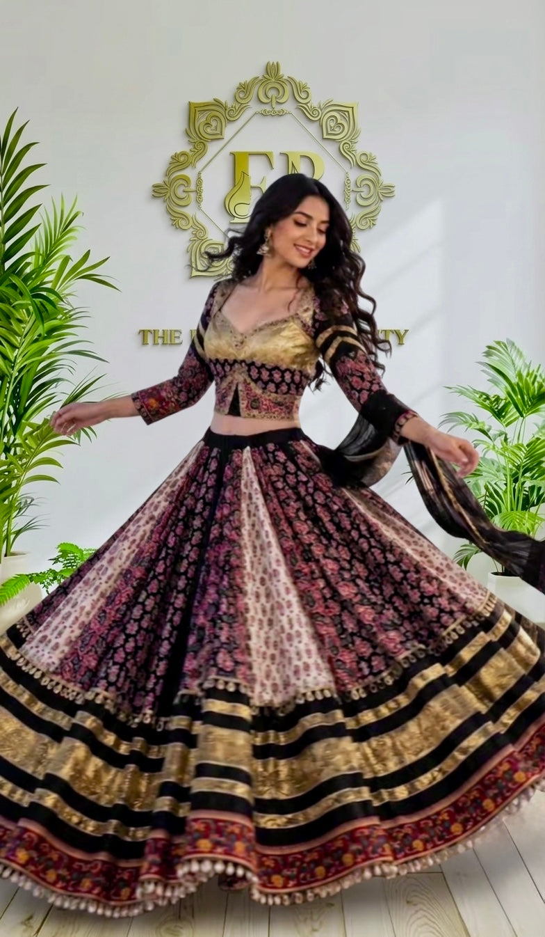 A woman modeling a traditional Indian lehenga choli set, featuring a gold and black embroidered blouse with a heavily patterned, flared skirt in black, gold, and pink. She is posing in front of a white wall with a gold logo and green plants.