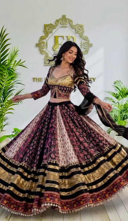 A woman modeling a traditional Indian lehenga choli set, featuring a gold and black embroidered blouse with a heavily patterned, flared skirt in black, gold, and pink. She is posing in front of a white wall with a gold logo and green plants.
