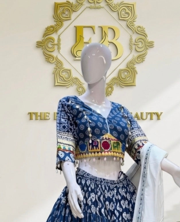 A blue and white printed traditional outfit displayed on a mannequin, featuring a patterned blouse with colorful embroidered details at the hem and sleeves, a matching printed skirt, and a white draped scarf. The background shows a gold decorative logo and the text 'THE BEAUTY'.