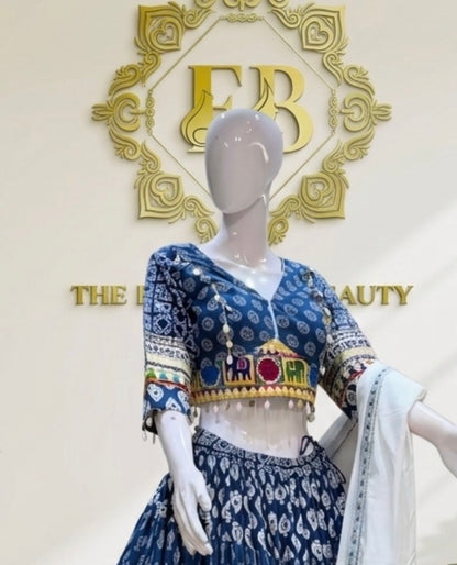 A blue and white printed traditional outfit displayed on a mannequin, featuring a patterned blouse with colorful embroidered details at the hem and sleeves, a matching printed skirt, and a white draped scarf. The background shows a gold decorative logo and the text 'THE BEAUTY'.