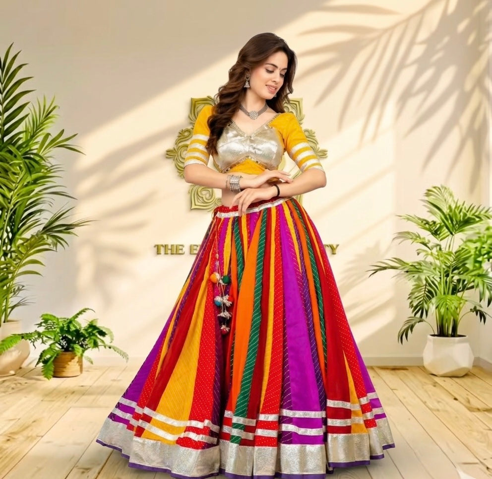 A woman models a vibrant Indian ethnic wear set consisting of a yellow and silver embroidered blouse and a highly colorful, striped skirt with purple, red, orange, green, and yellow panels, set against a warm beige wall with decorative plants.