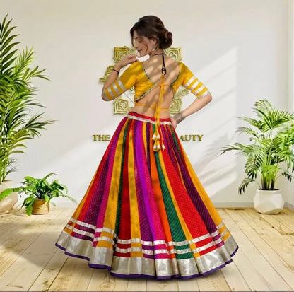 A woman models a vibrant, multicolored traditional Indian lehenga choli set in a brightly lit room with potted plants. The outfit features a yellow sleeveless blouse with gold trim and a highly flared skirt with vertical stripes of red, orange, yellow, green, pink, and purple, finished with a metallic silver border.