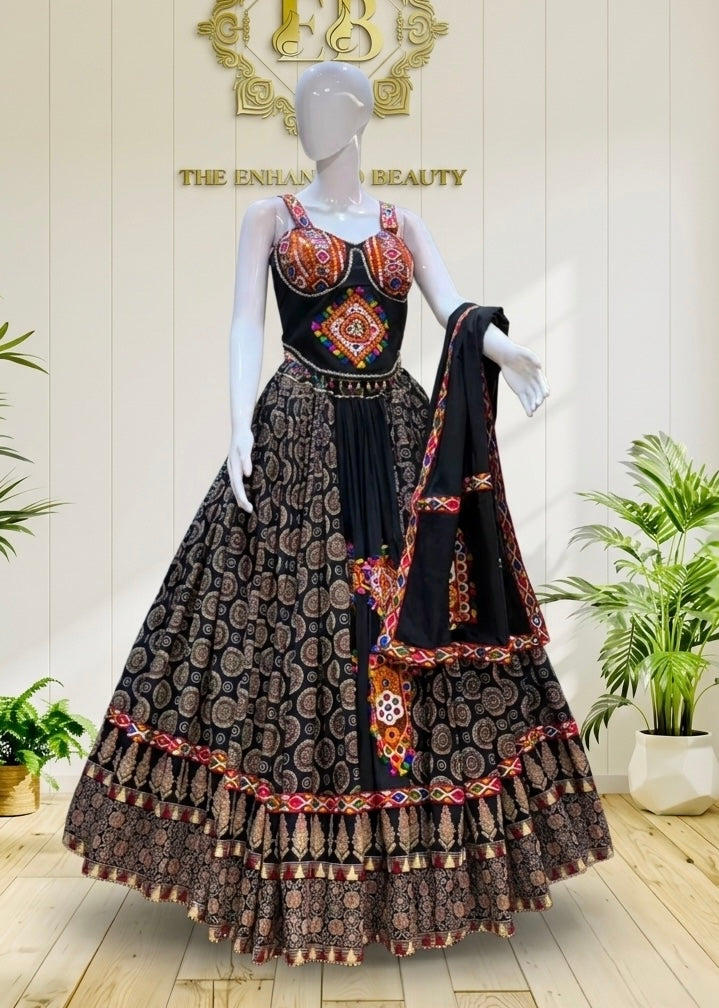 A black and brown patterned traditional Indian dress displayed on a white mannequin, featuring a heavily embroidered black bodice with red, orange, and gold details, a matching patterned skirt, and a black dupatta draped over one arm, set against a light wood-paneled wall with decorative plants.