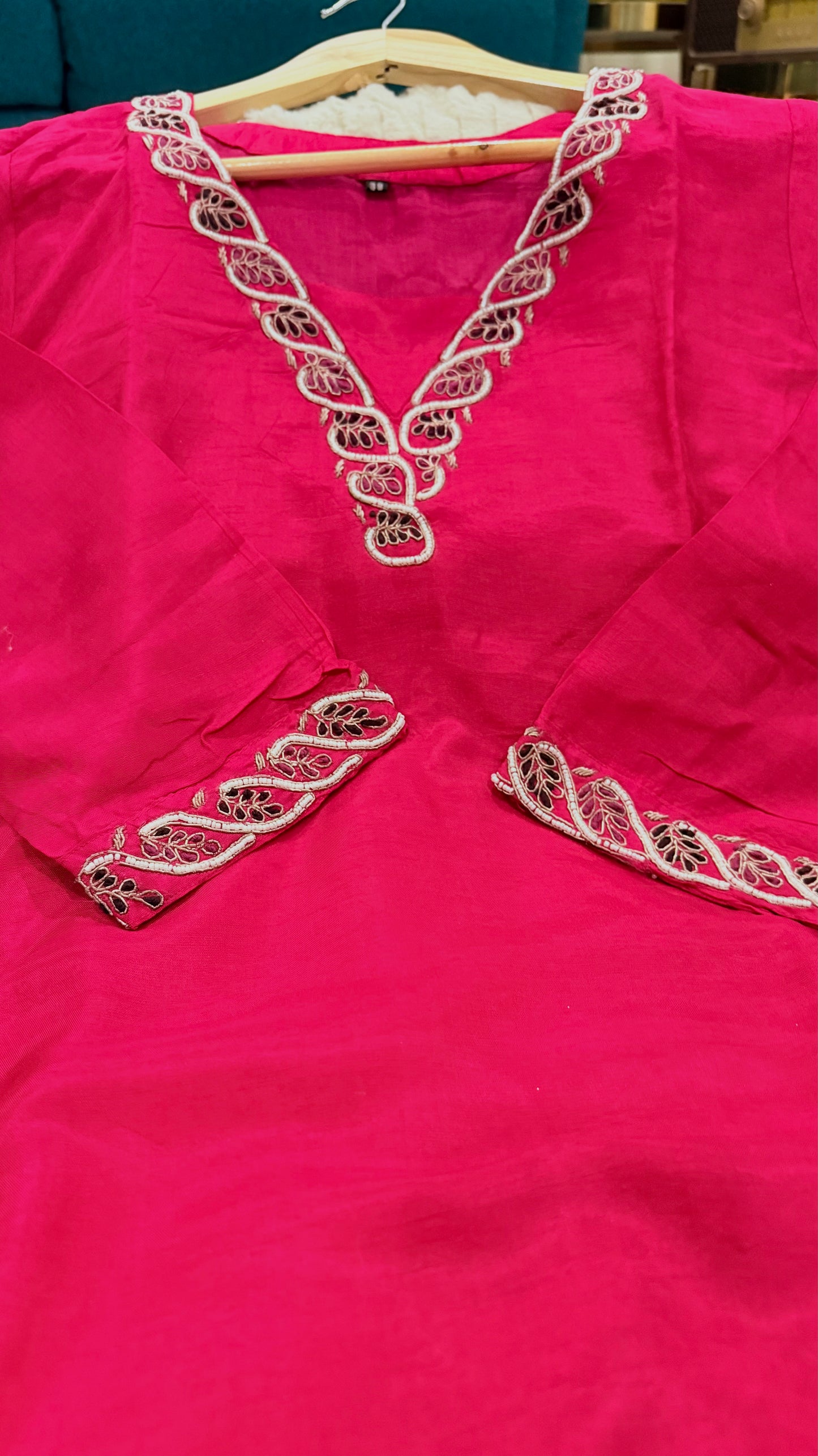 A bright pink kurta or tunic hanging on a wooden hanger, featuring ornate white and metallic embroidery around the V-neckline and sleeve cuffs.