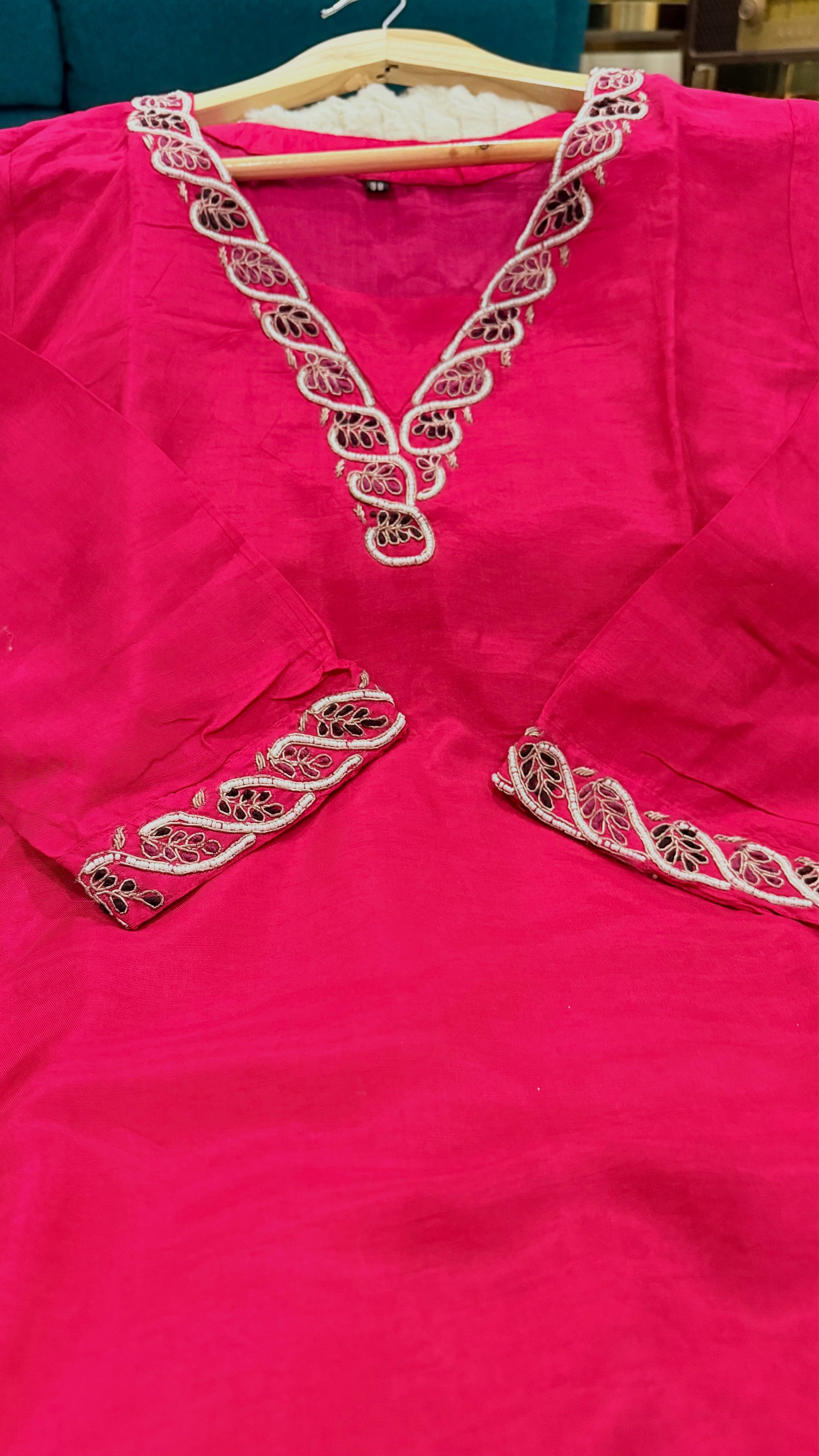 A bright pink kurta or tunic hanging on a wooden hanger, featuring ornate white and metallic embroidery around the V-neckline and sleeve cuffs.