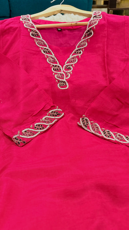 A bright pink kurta or tunic hanging on a wooden hanger, featuring ornate white and metallic embroidery around the V-neckline and sleeve cuffs.