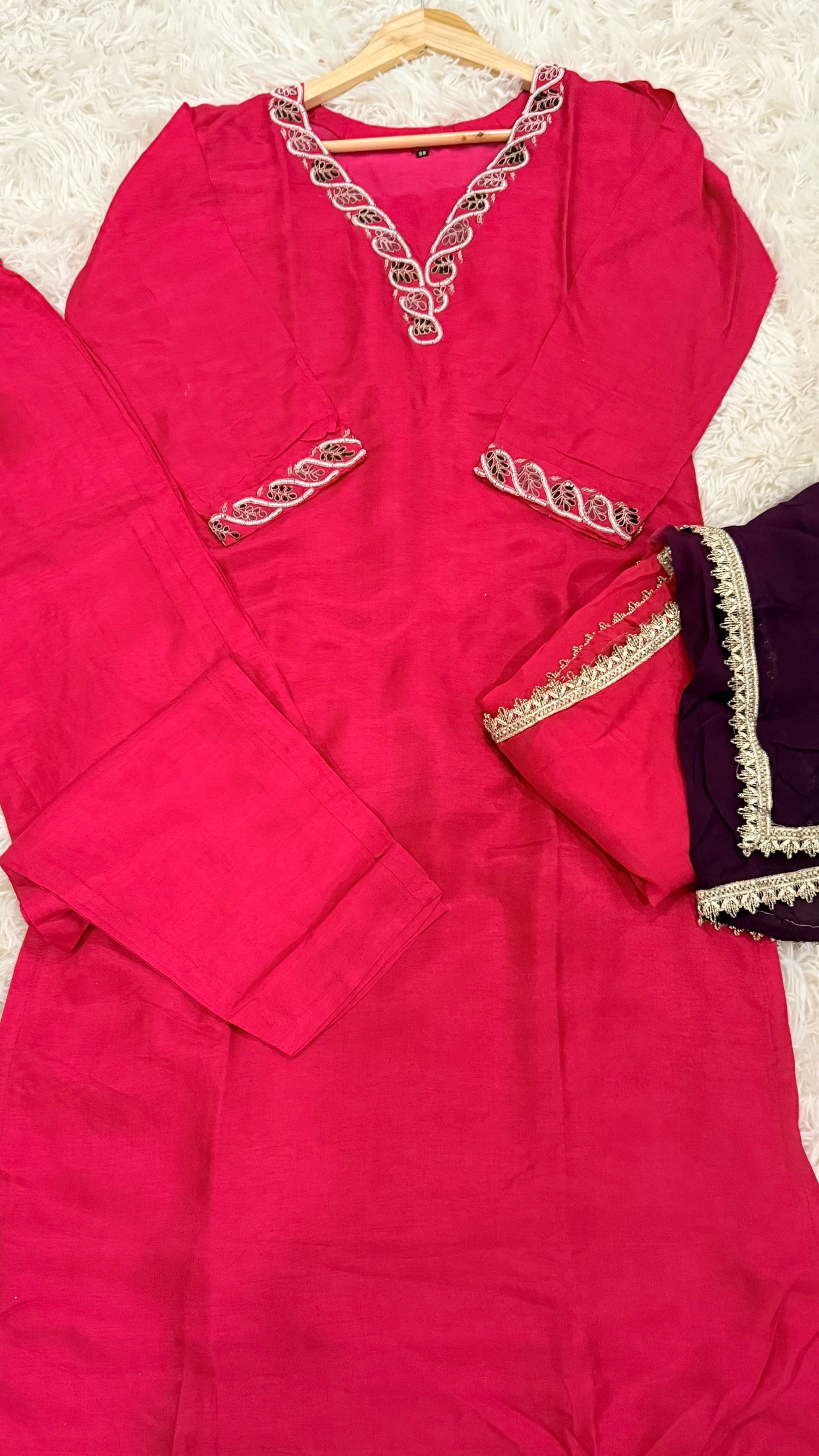 A bright pink traditional kurta set laid flat on a white textured rug, featuring a pink kurta with silver embroidered details on the neckline and chest, matching pink pants, and a folded dark purple dupatta with gold border trim.