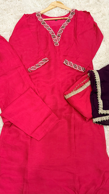 A bright pink traditional kurta set laid flat on a white textured rug, featuring a pink kurta with silver embroidered details on the neckline and chest, matching pink pants, and a folded dark purple dupatta with gold border trim.