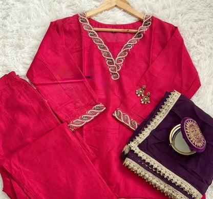 A traditional Indian outfit set featuring a bright pink kurta and matching trousers, styled with a folded dark purple dupatta decorated with gold zari embroidery and a small round mirror, laid out on a white faux fur background.