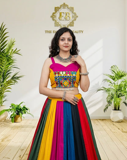 A woman modeling a vibrant, multicolored traditional Indian lehenga choli dress. The dress features a bright pink and yellow embroidered bodice with a multicolored striped skirt in red, orange, yellow, green, blue, and black.