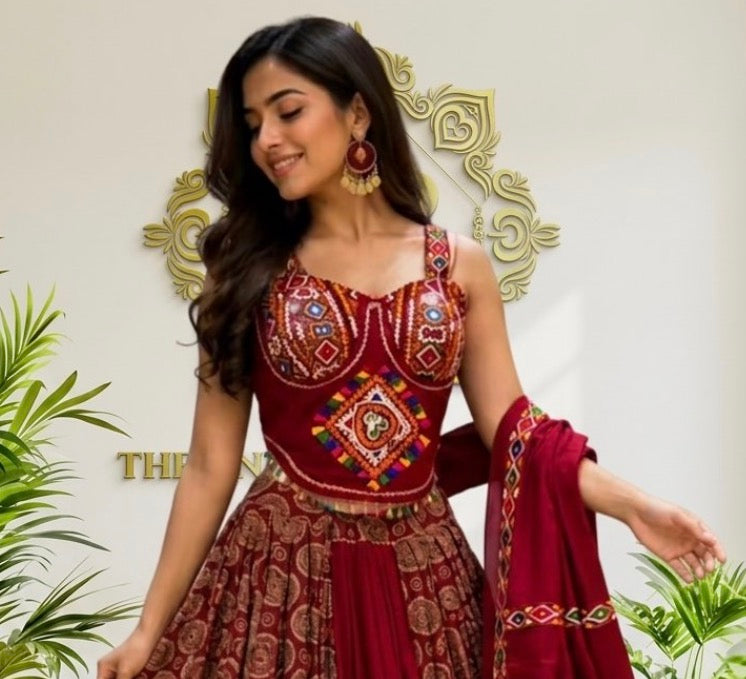 A woman modeling a traditional Indian red lehenga choli set with intricate gold and multicolor embroidery, standing in front of a decorative gold wall panel with green plants on either side.