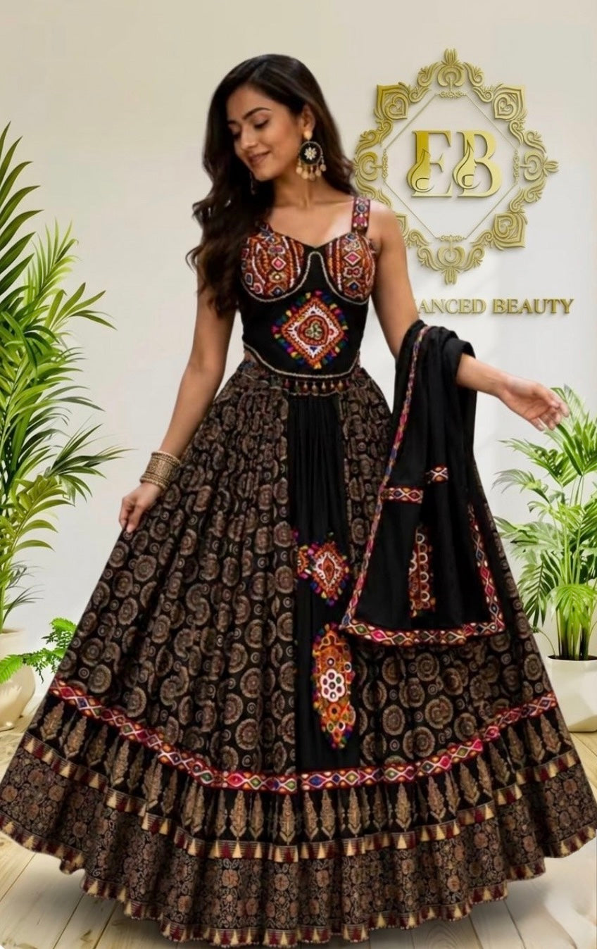A woman models a traditional Indian ethnic wear set featuring a black and brown printed lehenga choli with intricate red and gold embroidery, paired with a black dupatta. The background is a clean indoor setting with a gold 'FB Advanced Beauty' logo and potted plants.