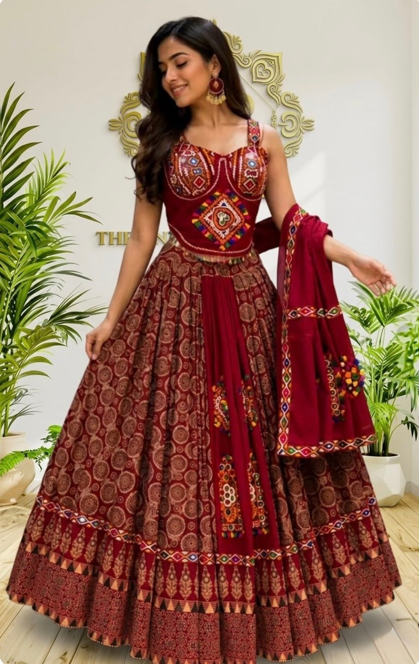 A woman models a traditional Indian lehenga choli set in deep red and maroon, featuring intricate gold and multicolor embroidery, a flared patterned skirt, and a matching dupatta, set against an indoor background with decorative plants.