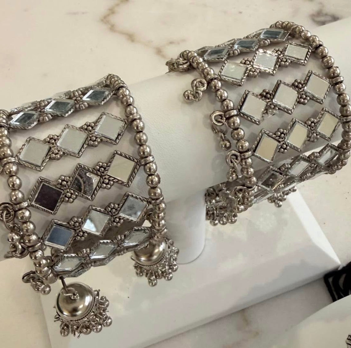 A pair of wide silver-toned bangles with mirrored square tiles and dangling jhumka-style charms, displayed on a white jewelry stand.