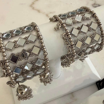 A pair of wide silver-toned bangles with mirrored square tiles and dangling jhumka-style charms, displayed on a white jewelry stand.
