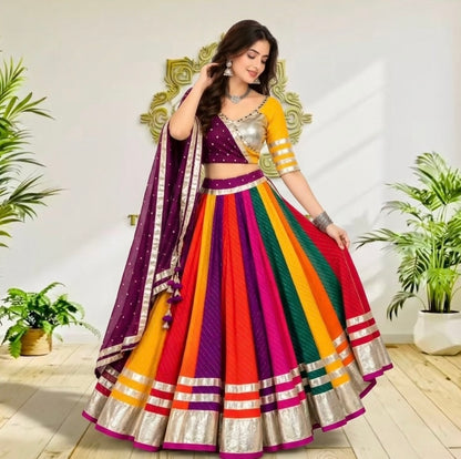 A woman models a vibrant, multicolored traditional Indian lehenga choli set in a bright indoor setting with potted plants. The outfit features a purple choli with gold embroidery, a multicolor striped skirt (yellow, orange, red, green, pink), and a matching purple dupatta with gold borders.