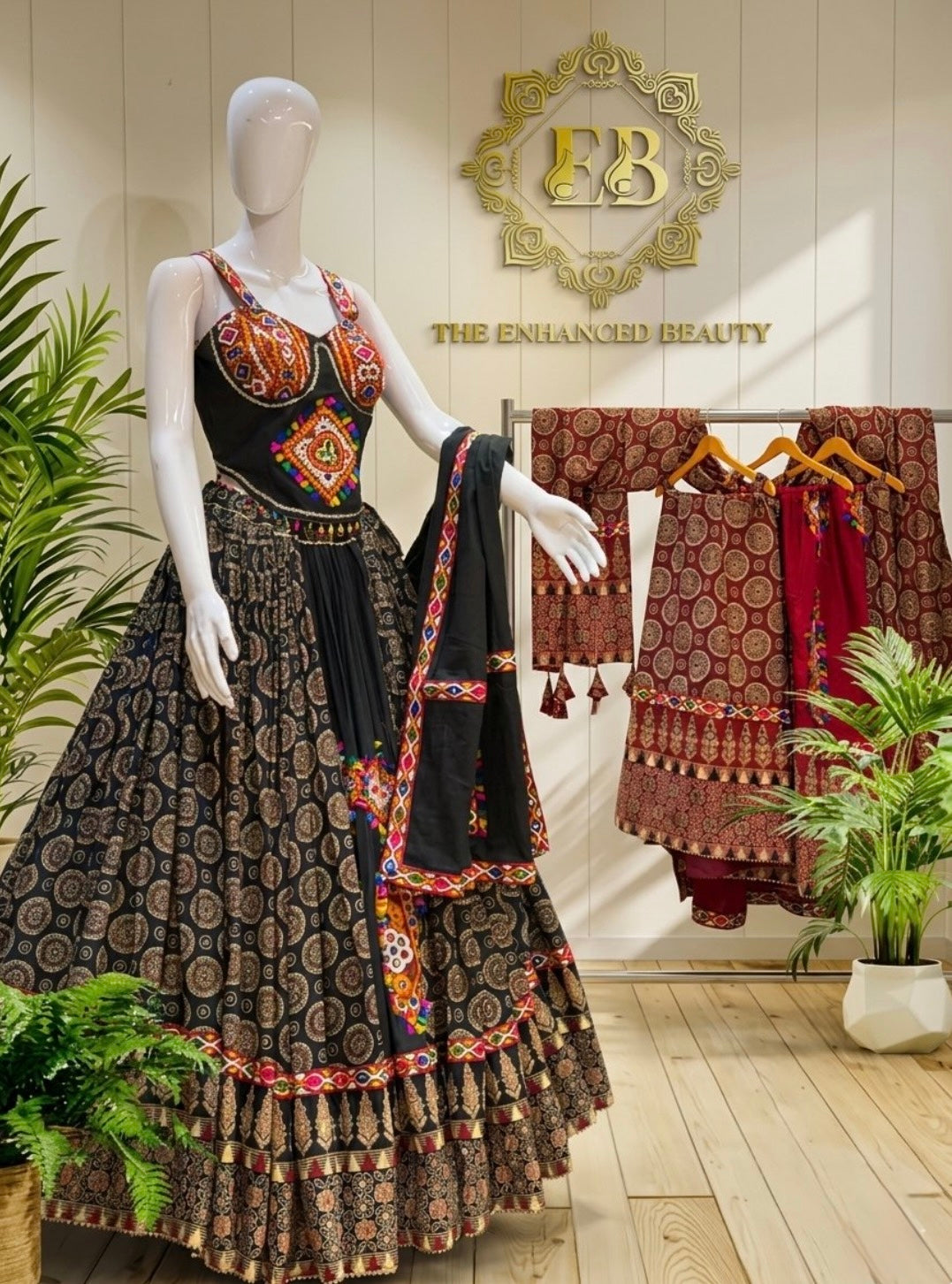 A black Indian ethnic lehenga choli set with intricate red, gold, and orange embroidery is displayed on a white mannequin in a boutique setting. The background features a wooden wall with a gold 'EB' logo, a clothing rack with matching garments, and potted green plants.