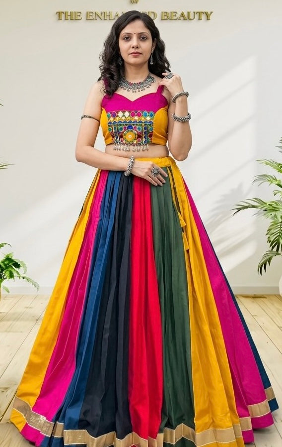 A woman models a vibrant, multicolored traditional Indian outfit consisting of a sleeveless crop top with colorful embroidery and a matching flared skirt with vertical stripes of yellow, pink, red, navy, and green, set against a light beige wall with decorative plants.