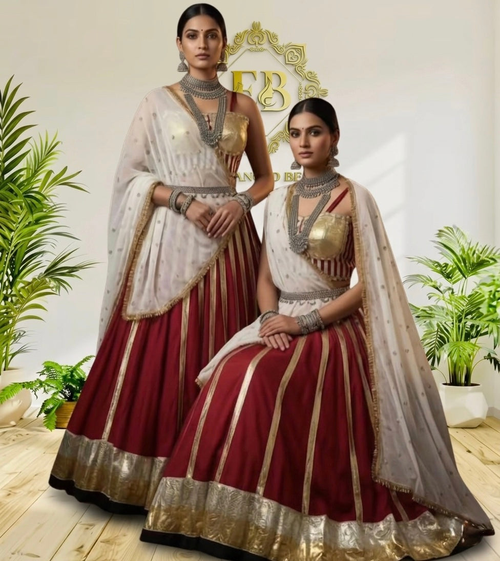 Two women modeling traditional Indian ethnic wear, featuring deep red lehengas with gold zari stripes and white dupattas, posed in front of a decorative wall with plants.