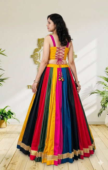 A woman wearing a vibrant, multicolored traditional Indian lehenga choli dress, shown from the back in an indoor setting with a light-colored wall and wooden floor.